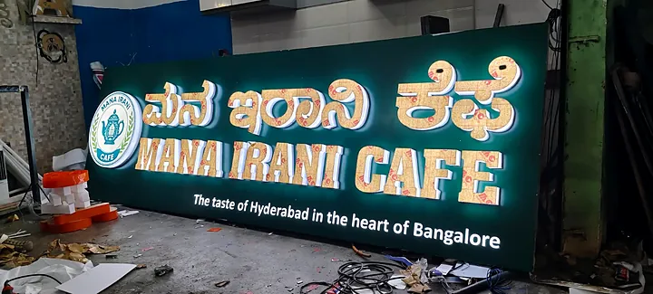 Mana Irani Cafe board lit up in the workshop: gold letters on green
